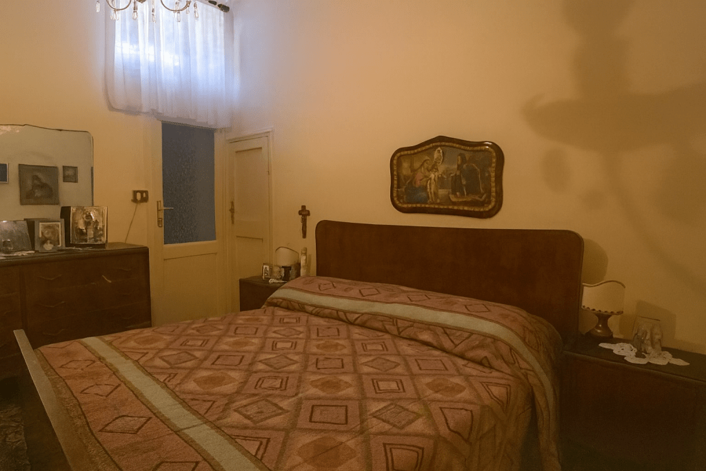 Camera da letto datata con scarsa illuminazione, arredi tradizionali scuri e oggetti religiosi.