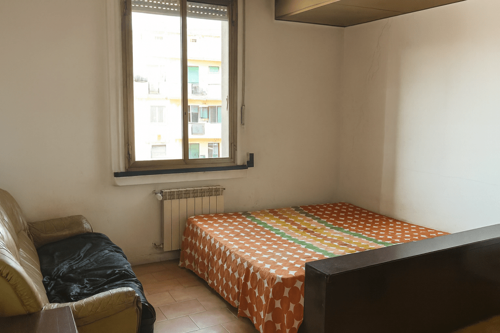 Una piccola stanza da letto datata con pareti bianche, un letto singolo con copriletto a righe, un calorifero sotto una finestra con infissi vecchi e un divano sulla sinistra.