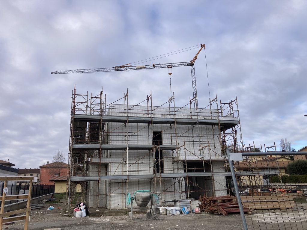 Vista di un cantiere edile disordinato con cielo nuvoloso, ponteggi, gru, reti arancioni e detriti metallici sparsi sul terreno.
