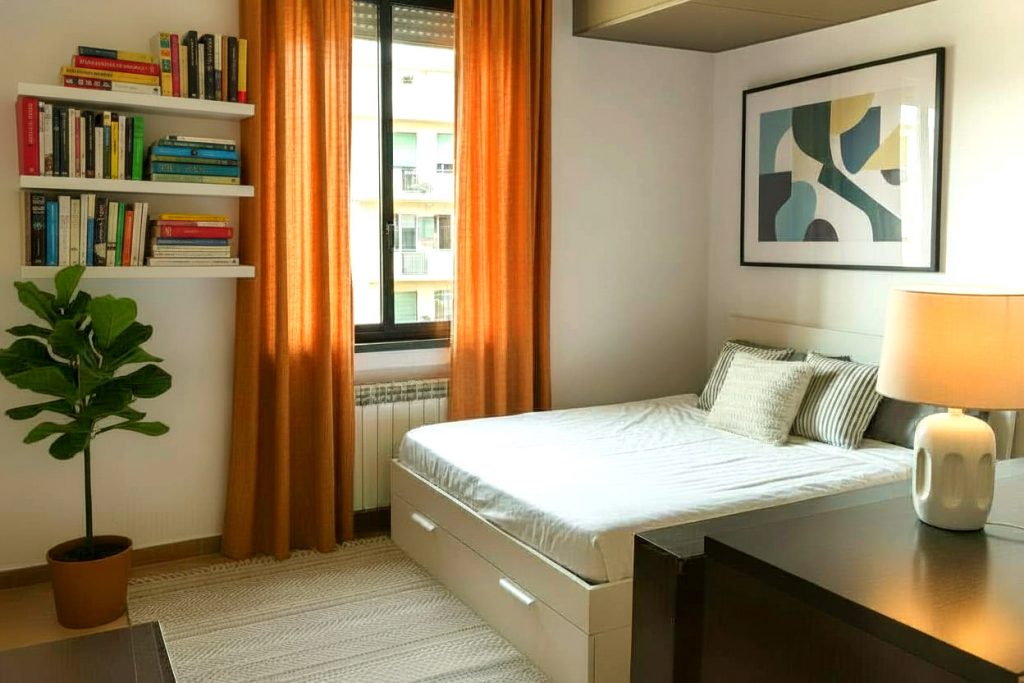 Una moderna camera da letto ristrutturata con letto matrimoniale, biancheria bianca, tende arancioni alla finestra con nuovi infissi, mensole con libri, una pianta ornamentale, una lampada da tavolo e un quadro astratto sulla parete.