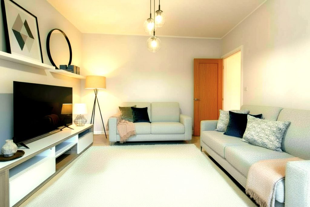 Soggiorno moderno e luminoso con pareti grigio chiaro, divani chiari, cuscini decorativi, tappeto chiaro e lampade di design. La TV è su un mobile elegante, e sono presenti quadri e uno specchio rotondo. L'ambiente è fresco e accogliente.