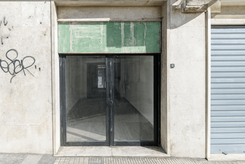 Facciata di un negozio vuoto con porta a vetri scura, mura esterne in cemento sporche con graffiti e un pannello superiore verde deteriorato, serranda metallica accanto.