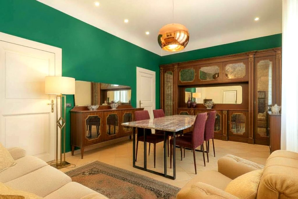 Salotto e zona pranzo modernizzati con pareti verde smeraldo, boiserie in legno scuro, tavolo da pranzo in marmo con base geometrica, sedie in velluto borgogna, lampada a sospensione Tom Dixon Melt e divani modulari in tessuto bouclé.