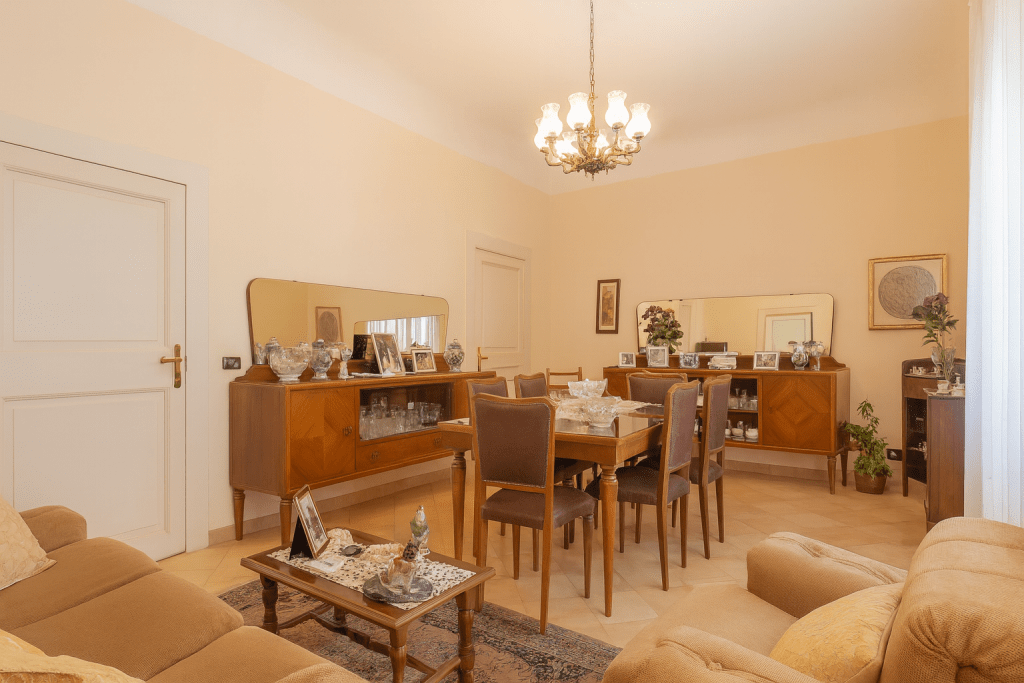 Salotto con zona pranzo arredato in stile classico con mobili in legno scuro, lampadario a candelabro, divani in tessuto chiaro e pareti color crema.