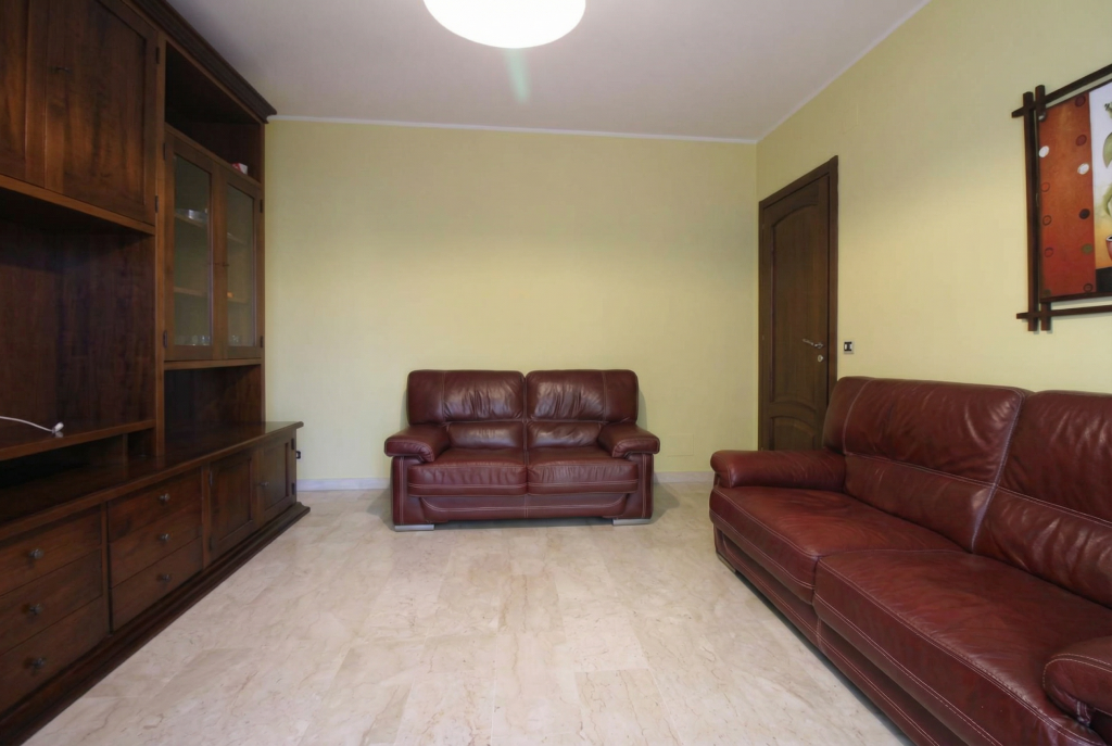 Soggiorno con pareti gialle, divano in pelle marrone datato, scaffali bianchi con piante e una TV su un mobile bianco. Illuminazione spenta e atmosfera poco accogliente.