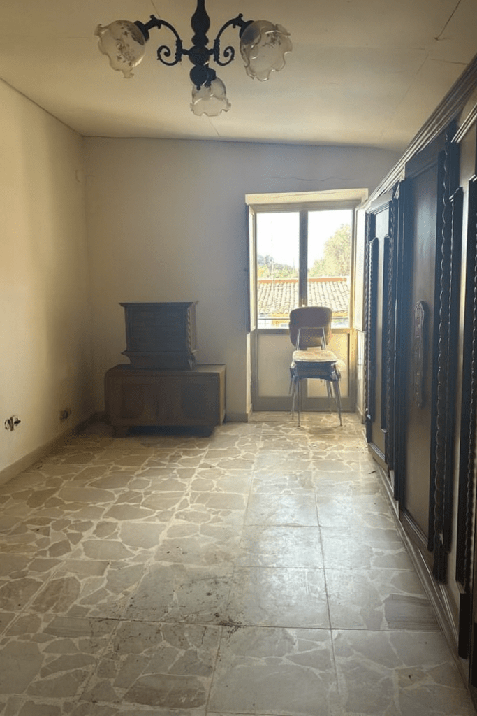 Stanza vuota e datata con pavimento a piastrelle antiche, pareti spoglie, lampadario obsoleto, finestra semplice, mobiletti scuri e armadio a muro ingombrante.