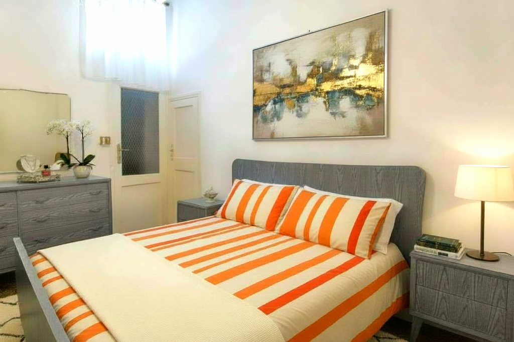 Camera da letto moderna e luminosa con arredi grigi, letto con lenzuola a strisce arancioni e quadro astratto.